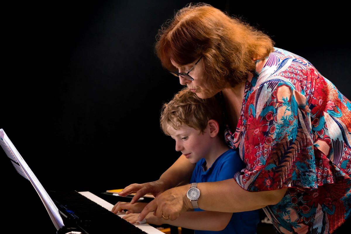 Professeure de piano d'Améthyste se tenant derrière son jeune élève, tout en se penchant vers lui pour l'aider à jouer du piano, illustrant un soutien pédagogique attentif.